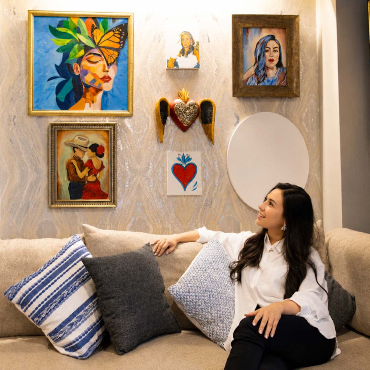 A woman sits on a beige sofa in a living room, looking at framed wall art. The room features multiple colorful paintings, decorative pillows, and a textured wall.