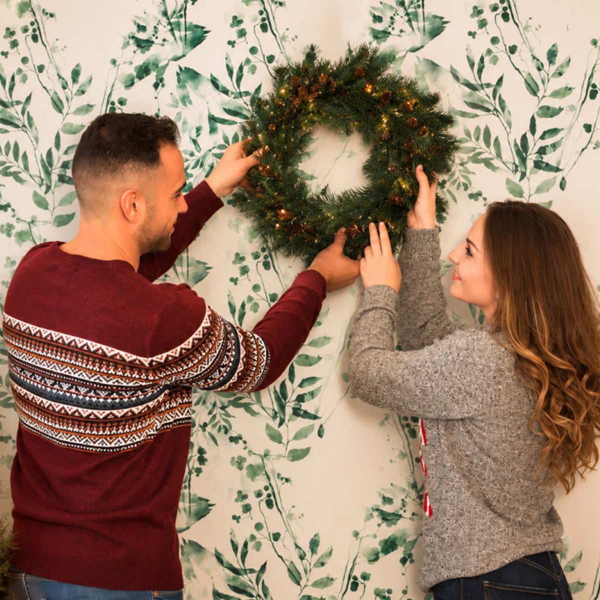 A man and a woman are hanging a green Christmas wreath with small warm lights on a white wallpapered wall with a green botanical pattern.