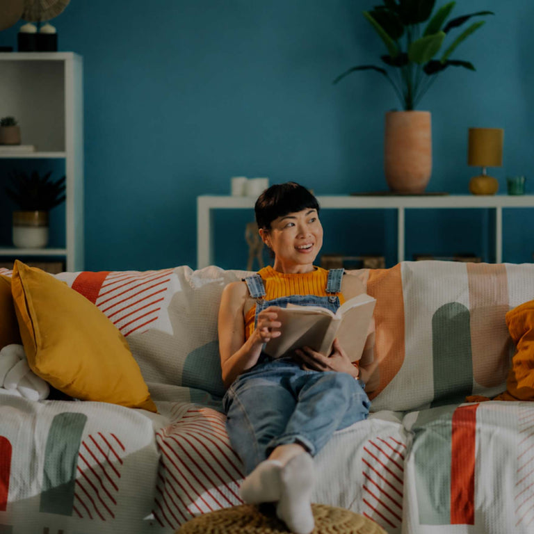 A woman sits on a patterned couch in a living room, reading a book. The room has a blue wall, white shelving, plants, and yellow and orange accent pillows.