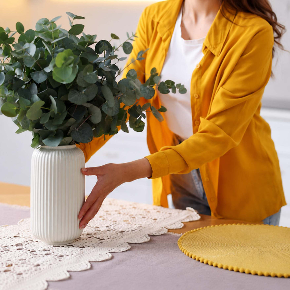 A woman in a yellow shirt and white top holds a white ribbed ceramic vase filled with green eucalyptus branches on a table with a lace doily and a yellow placemat.