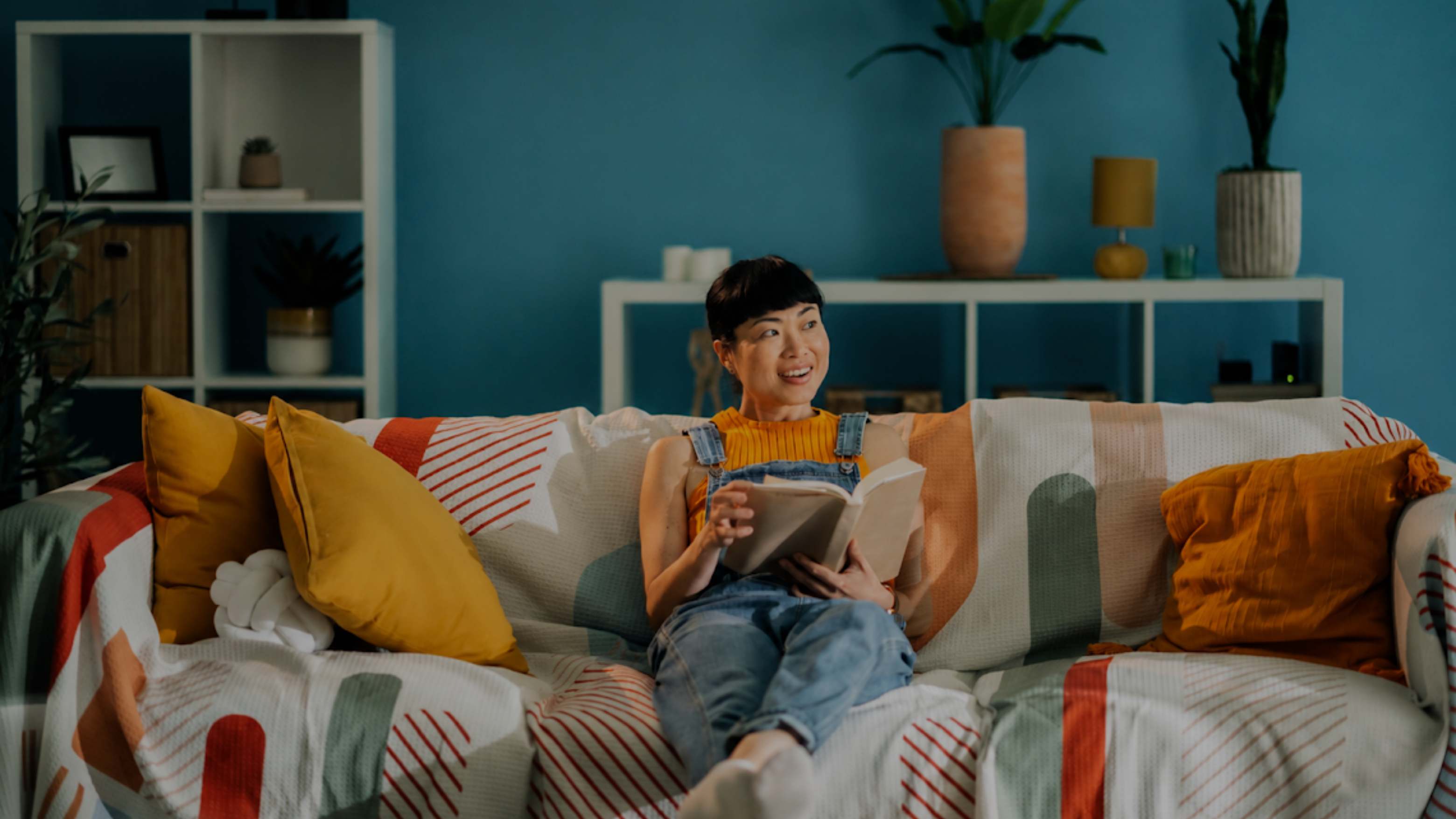 A woman sits on a patterned couch in a living room, reading a book. The room has a blue wall, white shelving, plants, and yellow and orange accent pillows.