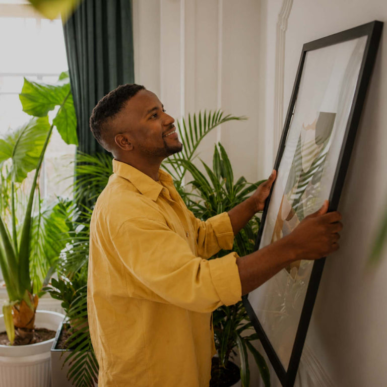 A man in a yellow shirt hangs a large framed picture on a white wall in a bright room with green plants.
